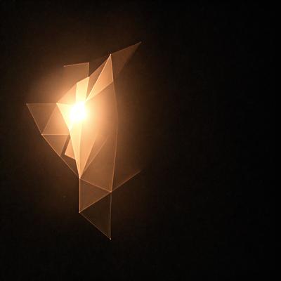 The primary subject of the image is a light source, possibly a light bulb or a sun, shining through a patterned or abstract shape. The setting is a dark background, which contrasts with the light source and the patterned shape. The visual style of the image is a combination of photo and digital art, as it appears to be a close-up of the light source with a unique pattern or shape in the foreground. The colors and mood of the image are predominantly dark, with the light source providing a contrast